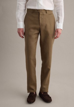 Man die bruine op maat gemaakte broek, wit overhemd met lange mouwen en donkerbruine suède loafers draagt, staande tegen een neutrale achtergrond.