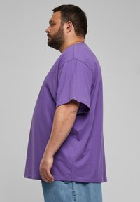 Tricou din bumbac violet cu mâneci scurte, croială lejeră și guler rotund. Asortat cu blugi din denim albaștri. Vedere laterală care arată lungimea și forma tricoului.