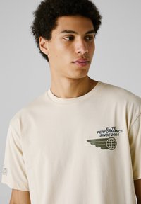Camiseta de algodón beige con un gráfico verde que presenta alas y un globo, y el texto "ELITE PERFORMANCE SINCE 2004" en el lado izquierdo del pecho. Corte clásico.