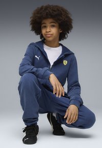 Tuta zip-up blu navy realizzata in morbido tessuto, con logo Puma ed emblema Ferrari. Scarpe sportive nere completano l'outfit.