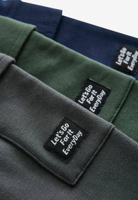 Drie gevouwen t-shirts in donkergrijs, olijfgroen en marineblauw, elk met een zwart label waarop staat "Laten We Elke Dag Gaan!"