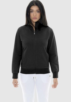 Donna con capelli lunghi e scuri che indossa una giacca nera con cerniera e pantaloni bianchi, in piedi davanti a uno sfondo chiaro e uniforme.