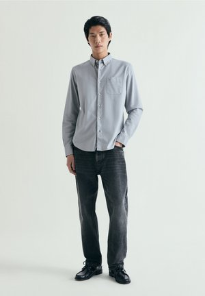 Bărbat tânăr în picioare purtând cămașă gri deschis cu nasturi, blugi închiși la culoare și pantofi negri, pe un fundal simplu, deschis la culoare.