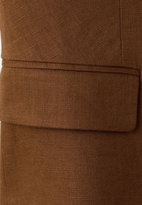 Tissu marron texturé avec un détail de poche à rabat horizontale sur un vêtement, montrant un tissage serré et des lignes de couture propres.
