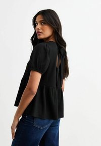 Blusa nera con texture, maniche corte a sbuffo, chiusura con laccetto sul retro e orlo a campana. Abbinata a jeans blu scuro.