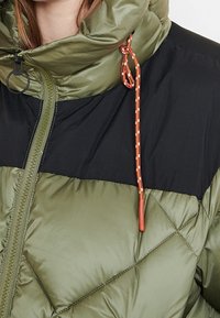Giacca puffer con una combinazione di colori verde e nero. Presenta un colletto alto, una texture trapuntata diagonale e un cordoncino arancione. Chiusura con zip.