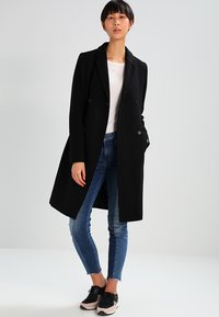 Sort dobbelbrystet frakke med skræddersyet pasform, båret over en hvid top, samt kombineret med slidte blå jeans og sorte sneakers.