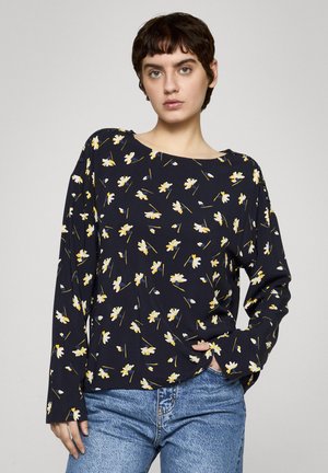 Junge Person mit kurzen dunklen Haaren, die ein marineblaues Langarmshirt mit gelbem und weißem Blumenmuster und hellblaue Jeans trägt.