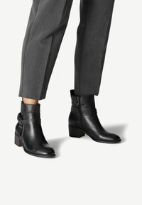 Botines de cuero negro con punta ligeramente afilada, cremallera lateral y un pequeño tacón apilado, que cuentan con un detalle de correa ajustable.