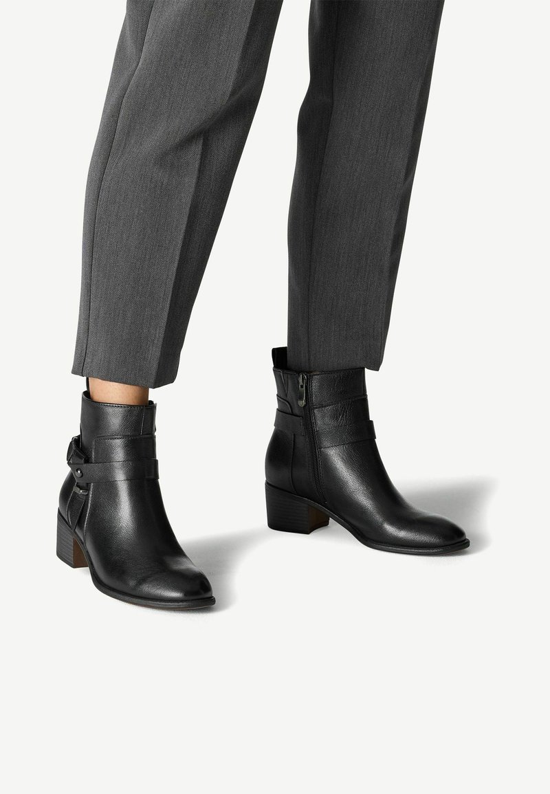Botines de cuero negro con punta ligeramente afilada, cremallera lateral y un pequeño tacón apilado, que cuentan con un detalle de correa ajustable.