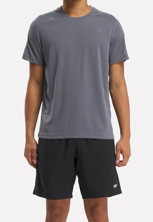 Hombre vistiendo una camiseta deportiva de manga corta gris y pantalones cortos deportivos negros, de pie con los brazos relajados a los costados.