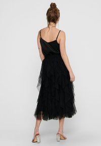 Schwarzes Satin-Camisole mit dünnen Trägern, kombiniert mit einem geschichteten schwarzen Tüllrock. Der Rock hat gestufte Rüschen und einen taillierten Bund. Silbern glänzende Absatzsandalen.