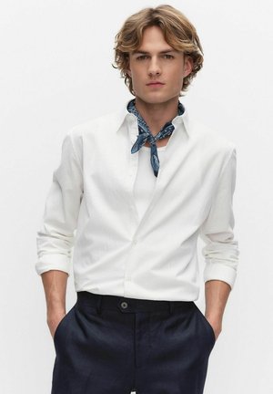 Młody mężczyzna w białej teksturowanej koszuli, niebieskiej wzorzystej apaszce na szyi i ciemnych spodniach, stojący z rękami w kieszeniach.