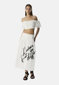 Witte cropped top met pofmouwen, gecombineerd met een lange witte rok met gedurfde zwarte tekst. Outfits aangevuld met zwarte sandalen.