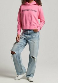 Růžová mikina s grafikou "SUPERDRY", v kombinaci se světle modrými širokými džíny s odřeninami, které mají trhliny a roztrhané okraje. Bílé tenisky.