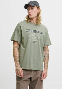 Olivengrøn bomulds t-shirt med "ORIGINALS" tekst, korte ærmer, rund hals og en afslappet pasform. Har et grafisk design og stingdetaljer.