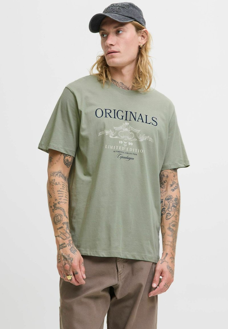 Olivengrøn bomulds t-shirt med "ORIGINALS" tekst, korte ærmer, rund hals og en afslappet pasform. Har et grafisk design og stingdetaljer.