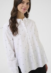 Chemise blanche à bouton avec un motif monogramme noir répétitif, manches longues, col pointu et ourlet arrondi. Fabriquée en tissu léger.