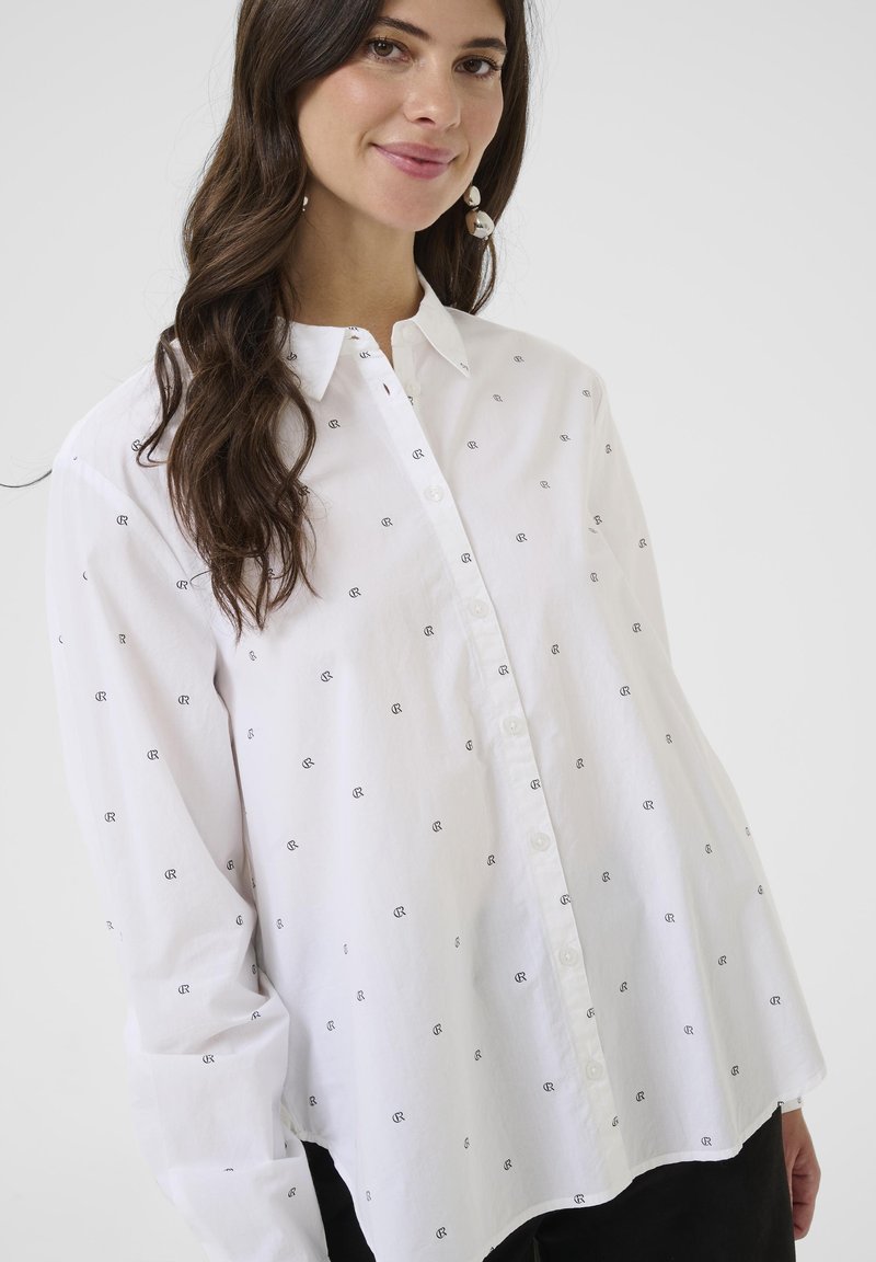 White button-up shirt featuring a lightweight fabric, embroidered black patterns, a pointed collar, and slightly oversized sleeves.