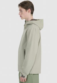 Young man in light beige hooded jacket with zippered pocket, standing sideways against plain light background.
