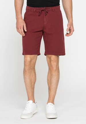 Uomo che indossa pantaloncini rossi con coulisse e scarpe da ginnastica bianche, in piedi davanti a uno sfondo chiaro uniforme con le mani rilassate ai lati.