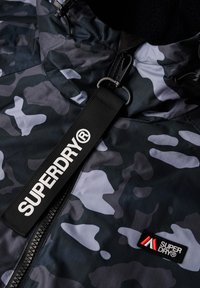 Czarna kurtka w kamuflażu z fakturowaną warstwą zewnętrzną, prążkowanym kołnierzem, pełnym zamkiem błyskawicznym oraz wyraźnym białym logo "SUPERDRY" na taśmie.