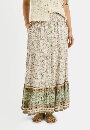 Falda larga con estampado floral y borde en verde y beige, combinada con un top de crochet crema con botones y sandalias doradas de punta abierta.