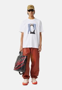 Biała bawełniana koszulka z nadrukiem graficznym, czerwone skórzane spodnie, czarna torba na ramię z czerwonym logo oraz białe sneakersy z pomarańczowymi akcentami.