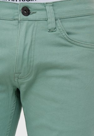 Denim de color verde claro y elástico, con una textura suave, que presenta un cierre de botón y bolsillos frontales, con costuras visibles.