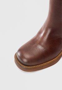 Καφέ δερμάτινο μποτάκι αστραγάλου με στρογγυλεμένη μύτη και ομαλή υφή. Διαθέτει αντίθετη ανοιχτή ραφή κατά μήκος της σόλας.