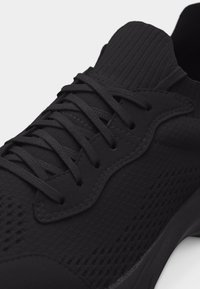 Chaussure de sport noire avec dessus en maille, surpiqûres et lacets croisés sur fond blanc.