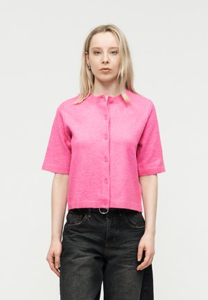 Jonge vrouw met steil blond haar, gekleed in een felroze kortmouwig truitje met knopen en een zwarte spijkerbroek, tegen een lichte achtergrond.