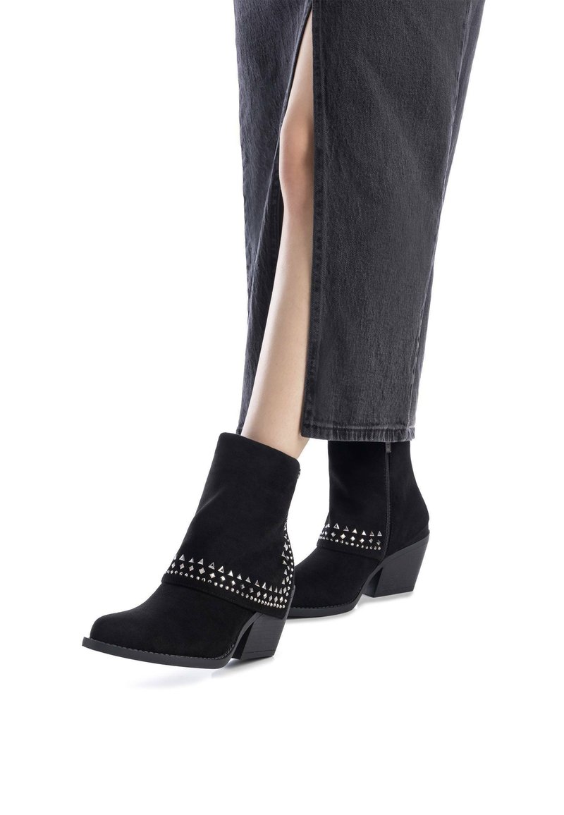 Botas negras hasta el tobillo de ante con tacón cuadrado, que presentan una banda de tachuelas alrededor de la parte superior. El diseño es elegante y aerodinámico.
