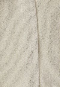 Stickat tyg i krämfärg med texturerad yta och små öppningar, som visar vågiga mönster och ett mjukt, lätt utseende.