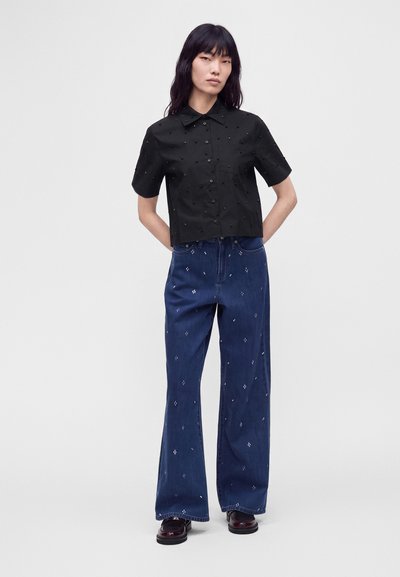 Femme debout, les mains derrière le dos, portant une chemise noire à manches courtes boutonnée et un jean large bleu avec de petits motifs blancs.