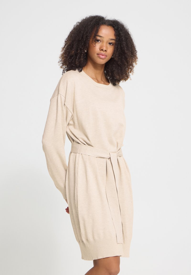 Jeune femme aux cheveux bouclés portant une robe tricotée beige à manches longues et ceinturée, debout devant un fond clair uni.