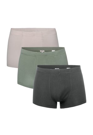 Muscular man wearing green boxer briefs, looking sideways with one hand adjusting waistband against plain white background.