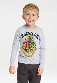 Gray long-sleeve t-shirt featuring a colorful Hogwarts crest design with "HOGWART'S" printed above it. Paired with dark brown pants.