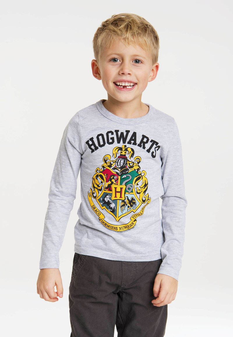 Gray long-sleeve t-shirt featuring a colorful Hogwarts crest design with "HOGWART'S" printed above it. Paired with dark brown pants.