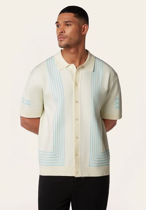 Man draagt een crème korte mouwen overhemd met lichtblauwe lijnpatronen, staat met zijn handen achter zijn rug en kijkt opzij.