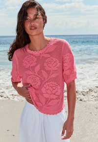 Femme debout sur une plage portant un haut rose en crochet avec des motifs de roses et un pantalon blanc ample, vagues de l’océan en arrière-plan.