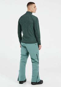 Langärmliges, dunkelgrünes Fleece-Oberteil mit hohem Kragen, kombiniert mit hellblauen wasserdichten Hosen mit Reißverschlüssen und schmal zulaufendem Bein-Design.