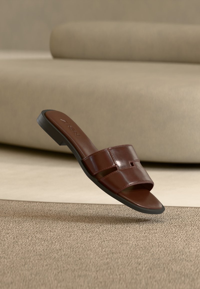Sandale à enfiler en cuir marron avec un design ajouré sur un tapis beige texturé devant un canapé de couleur neutre.