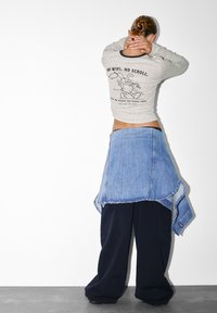 Šedá košeľa s dlhým rukávom, na ktorej je čierny text a ilustrácia na zadnej strane, spárovaná s vrstvenou modrou denimovou sukňou a voľnými čiernymi nohavicami.