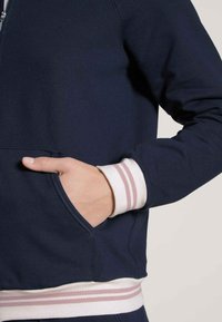 Marine blauwe trui met rits en een voorzak, met witte en roze gestreepte manchetten en een zachte stoftextuur. Heeft een aansluitend ontwerp met zichtbare stiksels.