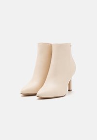 Bottines en cuir beige à bouts pointus et talons hauts fins, fermeture éclair latérale, présentées sur fond blanc.