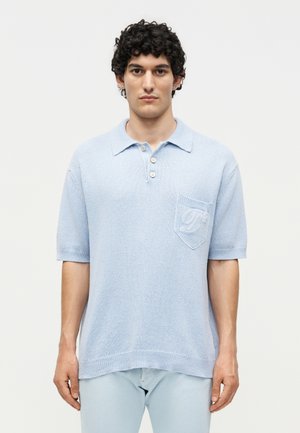 Άνδρας που φορά ανοιχτό μπλε πλεκτό πόλο με κοντά μανίκια, γιακά, τρία κουμπιά και τσέπη στο στήθος με κεντημένο γράμμα, στέκεται μπροστά σε λευκό φόντο.