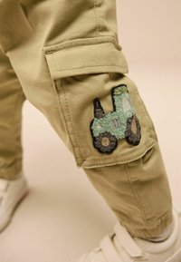 Beige cargo nadrág zöld traktor folttal a térdnél, törtfehér sportcipővel viselve, világos háttér előtt.