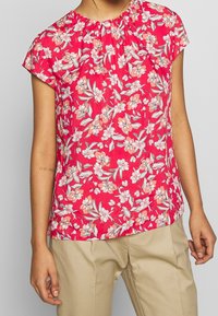 Bloemenblouse voor vrouwen in roze stof met korte mouwen en een geplooide halslijn. Het ontwerp bevat witte en koraalachtige bloemen met groene bladeren.