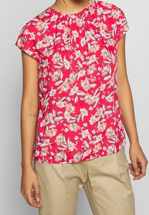 Blouse féminine fleurie en tissu rose, avec des manches courtes et un col froncé. Le design comprend des fleurs blanches et corail avec des feuilles vertes.